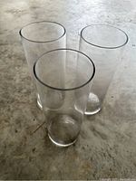 Three clear glass vases arranged together on a concrete floor, showing their cylindrical shape and clear glass material.