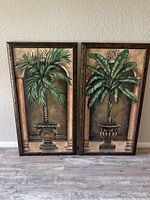Two palm tree canvas picture frames side by side on floor against wall showing full artwork and frames.