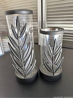 Two cylindrical glass candle holders with silver metal leaf designs, black base and rim, one taller and one shorter