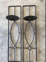 Photo showing two matching black metal wall candle stands placed flat on a concrete floor.