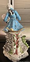 Lady figurine wearing blue coat with a bonnet and standing on a base decorated with tree and floral elements.