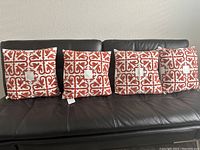 Four square decorative pillows with a red and white geometric pattern arranged on a black leather couch.