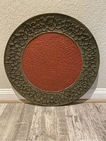 Front view of the decorative plate showing the floral patterned green border and red textured center on the floor against a wall.