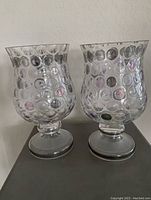 Photo shows two clear glass hurricane lamps with bubble pattern on the curved exterior bodies and round pedestal bases.