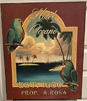 Photo of a rectangular painted wooden plaque featuring two green parrots perched on a beach scene archway with text.