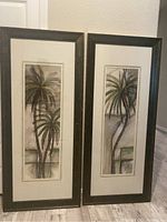 Two vertical picture frames side by side featuring stylized palm tree artwork with dark wood frames and light mats.