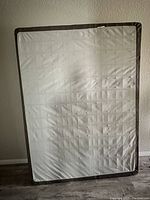 Full front view of the queen size box spring against a wall, showcasing the white quilted fabric surface with tufted pattern and brown trim edges.