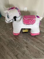 Electric white pony plush toy with colorful mane, pink saddle, and bridle, positioned next to a wall outlet on a wood floor.