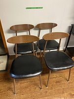 Four retro chairs with black vinyl seats and oval veneer backs, metal legs in brown finish, grouped together on wooden floor