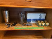 Shelf with Waterford crystal cocktail glasses in box, metal cocktail shaker, strainer, bottle openers, clear mixing glass, and ornate colorful shot glasses.