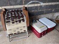 Four folded lawn chairs stacked side by side next to two coolers (Thermos and Coleman).