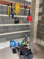 Wide shot of assorted car supplies including cleaning fluids, tools, jumper cables, and a collapsible bag on a concrete floor and a wall with mounted tools