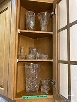 Wooden cabinet with three shelves containing assorted crystal decor: top shelf has two tall crystal vases, middle shelf has a small crystal candle holder, a medium bowl, and two cut glass pieces, bottom shelf has a tall vase and a small vase.