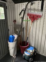 Garden tools including leaf rake with red head and wooden handle, large shovel, hoe, and other long-handled tools stored upright in containers in a shed corner.
