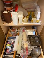Overview of assortment of kitchen bakeware and storage items including plastic containers, mixing bowls, rolling pins, and baking tins organized in cabinet shelves.