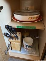 Full view of the kitchen items including CorningWare lidded frying pan on Rosenthal platter, knife block with knives, ceramic dish on marble base and lidded ceramic pot in wood cabinet.