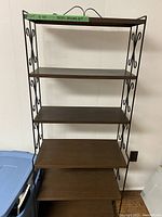Metal shelving unit with five faux wood shelves and black ornate sides against white wall. Shelves evenly spaced with decorative metal scrollwork sides visible.