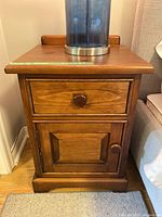 Front view of one wooden bedside table showing drawer and lower cabinet door with round knobs, and a lamp resting on top.