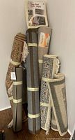 Several rolled rugs, runners, and mesh anti-slip pads leaning against a wall, showing different designs and labels.