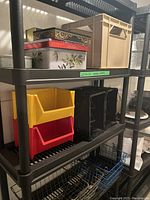 Black plastic shelving unit loaded with various storage containers including cookie tins, plastic milk crates, stacking bins and wire baskets.