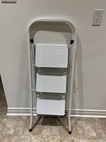 White metal step ladder folded and leaning against wall showing three ribbed steps