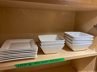Shelf displaying seven stacked small square plates, seven stacked medium square bowls, and additional larger square bowls all in white ceramic.