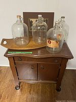 Full view of wooden cabinet holding 4 clear glass jugs on top, teak tray under jugs, sepia photo behind jugs