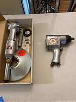 Photo showing an air powered 7P grinder with attached guard and handle in a box alongside an Alltrade 1/2 inch pneumatic impact wrench on a table.