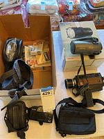 View of multiple cameras, black carrying case, and a box of accessories on a table with bedding in background