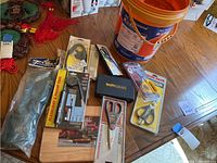 Photo showing lot contents on table including cutting board, various kitchen scissors and shears, chef's tool kit, flashlight and pickup tool set in packaging, and an orange bucket.