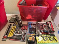 Various hand tools displayed on table including pliers, screwdrivers, drill bit sets, measuring tapes, and a red storage bin containing additional tools.