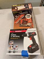Three boxed items stacked: First Alert Ready-Lite flashlight on top, Black & Decker Electric Brad Nailer in middle, Drill Master Cordless Drill on bottom.