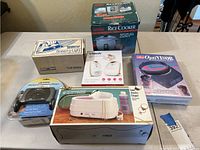 Collection seen on table includes boxed Coleman rice cooker, GPX cassette player in original package, Vtech baby monitors, Speed-o-Light iron in box, OptiVisor magnifier, and other household electronics items.