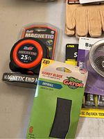 Items on display including a magnetic 25 ft measuring tape, drywall sanding block in packaging, and a pair of beige leather gloves.