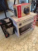 Photo showing the new Sunbeam 2-slice toaster, Sunbeam 6-slice toaster oven, and Pedrini paper towel holder all boxed and stacked on floor.