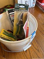 White garden bucket holding multiple pruning tools, mainly One Staff brand green handle pruners in packaging, yellow handle pruner also visible.