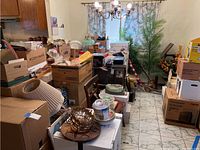 Room filled with boxes, kitchenware, artificial plants, and miscellaneous household items on tiled floor, chandelier visible but excluded