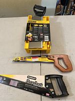 Overview of the Stanley clamping miter box with attached metal back saw on top, yellow plastic body and black handle. Below are two handsaws: Irwin with wooden handle and Stanley SharpTooth with black handle.