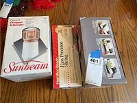 Photo showing Sunbeam Oskar Jr. Chopper & Grinder box, Hamilton Beach Easy Precision Slicing tool box, Chef's Choice electric sharpener box side by side on table.