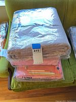 Top view of three blankets wrapped in plastic, folded and stacked on a green chair, showing label and brand Caprice on the pink blanket.