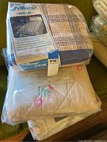 Image showing two bedspreads stacked, the top one in packaging labeled St. Marys Value-Line College Plaid, underneath a floral full-size bedspread in packaging.