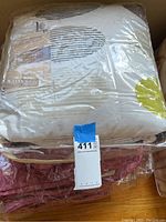 Two comforters stacked, one white with stripes and one reddish, both in plastic packaging.