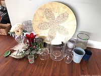 Wide composition showing the large decorative plate with textured flower pattern, multiple glass vases, candleholders and blue ceramic pots on wooden floor.