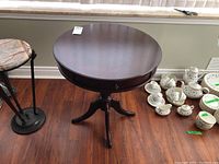 Round dark wood pedestal occasional table on 3 legs with a centered drawer, photographed from side angle, table surface, and open drawer view.