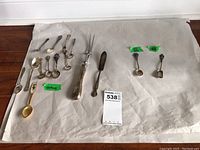 Full set of 12 sterling silver decorative spoons and forks laid out on white paper, showing variety of sizes and handle designs.