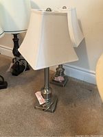 Two brushed nickel-colored table lamps with white fabric square bell-shaped shades on a carpeted floor near a wall.