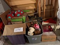Boxes and bins filled with party decor items including mushroom props, green fabric rolls, artificial flowers, ceramic bust, and glass decor pieces packed for transport.