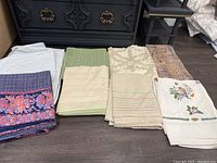 Multiple assorted rectangular table cloths folded and arranged on floor, showing variety of colors and patterns, including white, green, cream with floral and stripe designs, paisley, floral embroidery, and plaid floral border.