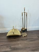 Brass fireplace tool set including a stand with three tools (poker, shovel, brush) and a brass ash scoop. All items are on a hardwood floor.