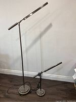 Photo showing one tall floor lamp and one short table lamp matching in design and finish, placed on dark floor against a light wall.